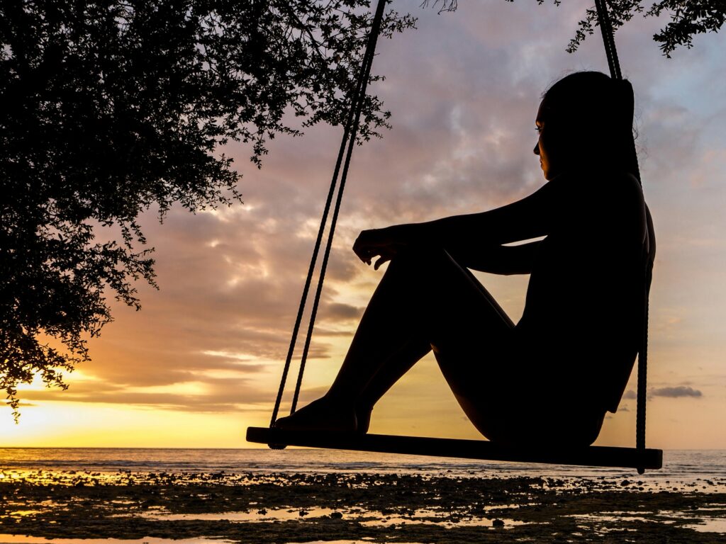 A serene sunset silhouette of a woman sitting on a swing by the seashore, evoking tranquility and reflection.